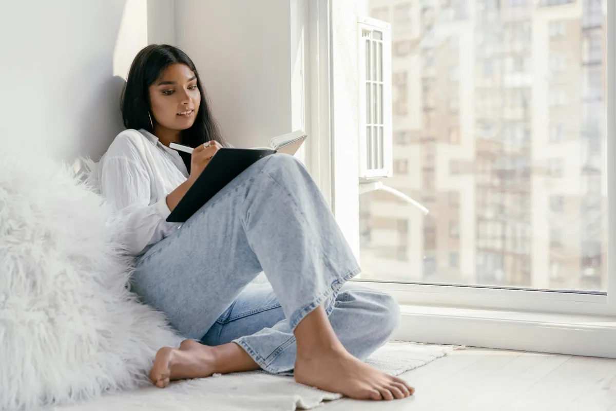 Persona escribiendo en un diario junto a una ventana con luz natural